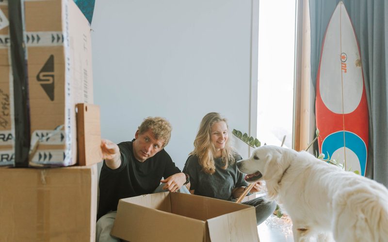 Casal desempacotando caixas de mudança no apartamento novo, com o cachorro por perto.